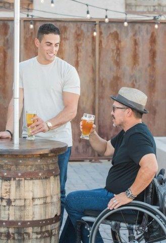 A man sitting in a wheelchair enjoying a glass of beer with his friend who is standing.