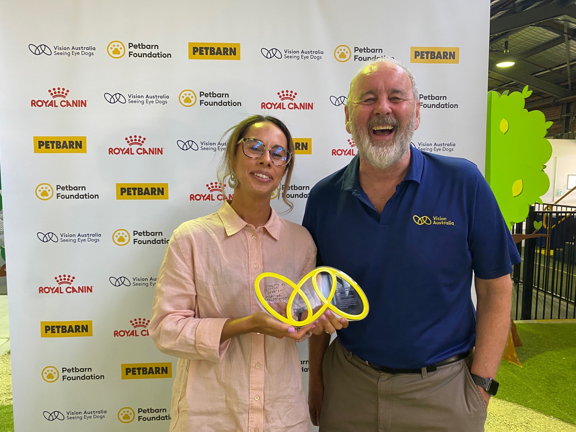 "Vision Australia CEO Ron Hooton and Petbarn Foundation manager Janelle Bloxsom smiling widely while Janelle holds the Vision Australia Award."